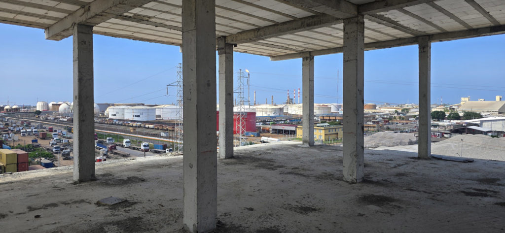 kama-gpl-hba-architecture-vue-interieure-niveau-superieur-zone-industrielle-vridi Vue depuis l'intérieur d'un niveau supérieur du bâtiment administratif KAMA GPL à Vridi — trame de colonnes béton cadrant la zone industrielle portuaire d'Abidjan avec réservoirs, voies ferrées et infrastructures logistiques